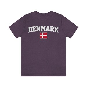 K&ouml;nnte beinhalten: Ein lila T-Shirt mit dem Wort "DENMARK" in wei&szlig;er, abgenutzter Schrift und der d&auml;nischen Flagge darunter.