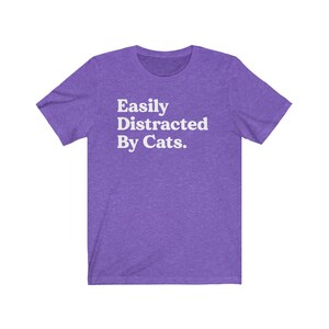 Może przedstawiać: Fioletowy T-shirt z kr&oacute;tkim rękawem z napisem "Easily Distracted By Cats." w kolorze białym.
