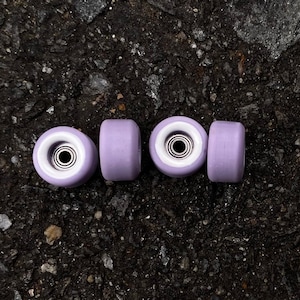 Puede incluir: Cuatro ruedas de skate de color morado con centros blancos y rodamientos negros.