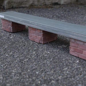 May include: A gray concrete bench with three brick supports. The bench is sitting on a gray asphalt surface.