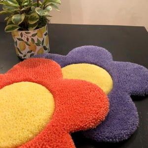 May include: Three flower-shaped floor mats in purple, orange, and yellow. The mats are made of a soft, fluffy material.