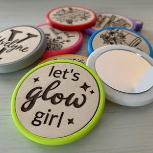 Peut inclure: Plusieurs miroirs compacts ronds en bois avec divers motifs et textes. L'un d'eux porte l'inscription "let's glow girl" en écriture noire. D'autres miroirs présentent des motifs floraux et des noms. Les miroirs ont des bords colorés.