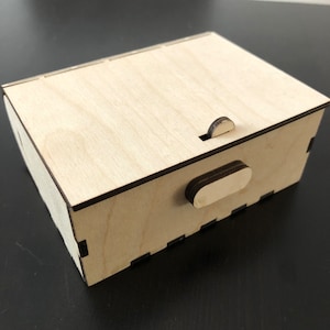 Peut inclure: Une petite boîte en bois avec un couvercle à charnière et un loquet coulissant. La boîte est en bois clair et a une finition naturelle.