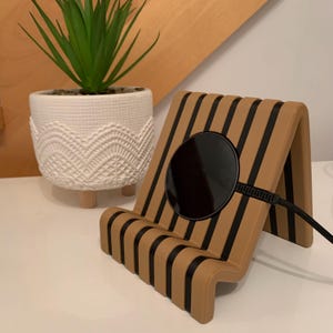 May include: A tan and black wireless phone charger with a striped design. The charger has a black charging pad and a black cord. A white ceramic pot with a green plant is in the background.