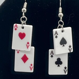 Puede incluir: Pendientes colgantes plateados con diseños de naipes. Cada pendiente presenta dos cartas rectangulares blancas con símbolos de corazones, diamantes, picas y tréboles rojos y negros, y la letra "A".