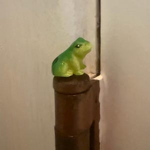 Pode incluir: Pequena estatueta de sapo verde com manchas pretas. O sapo está empoleirado em um objeto cilíndrico marrom. A estatueta tem um acabamento brilhante e está em um fundo neutro.