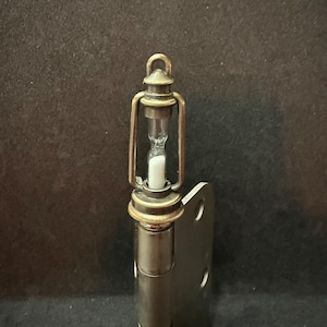 May include: A miniature hourglass encased in a bronze-colored lantern frame. The lantern is attached to a metal hinge. The hourglass contains white sand. The background is a dark, solid color.
