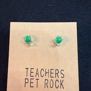 May include: Turtle-shaped stud earrings with green and silver detailing. The earrings are presented on a tan card with the text "TEACHERS PET ROCK" in black.