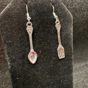 May include: Silver-toned spoon and fork earrings. Each earring features a miniature spoon and fork design, with intricate detailing. The earrings hang from silver-colored fish hook ear wires. The background is a dark, textured surface.