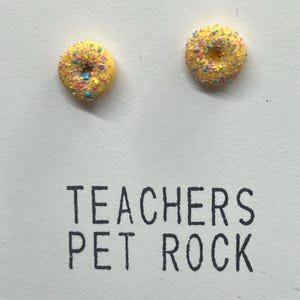 Puede incluir: Un par de pendientes amarillos en forma de donut con chispas de colores. Los pendientes están en una tarjeta blanca con el texto "TEACHERS PET ROCK".