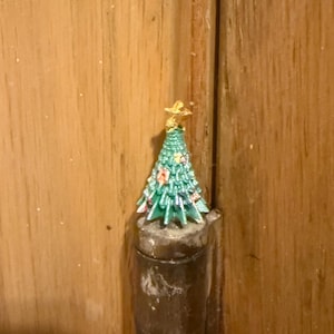 Puede incluir: Un pequeño árbol de Navidad en miniatura con una estrella dorada. El árbol es verde con detalles florales rosas y blancos. Se coloca sobre un objeto cilíndrico marrón oscuro, sobre un fondo de madera.