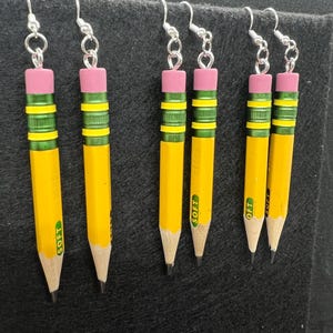 May include: Five pairs of yellow pencil earrings with pink erasers and green and yellow stripes. Each earring has a silver hook and the word "SOFT" printed on the pencil.