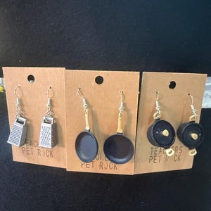 Könnte beinhalten: Ausgefallene Ohrringe auf braunen Karten. Die Ohrringe zeigen Miniatur-Reiben, Bratpfannen mit Bambusgriffen und schwarze Töpfe mit goldenen Knöpfen. Der Text "TEACHERS PET ROCK" ist auf einer Karte zu sehen.