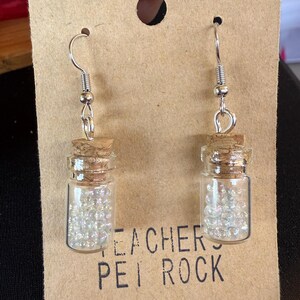 May include: A pair of dangle earrings featuring small glass vials filled with iridescent beads, each sealed with a cork stopper. The earrings hang from silver-colored fish hook ear wires. The earrings are displayed on a brown card with the text "TEACHERS PEI ROCK".