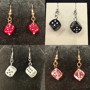 May include: A collection of dice-themed earrings. The earrings feature dice in various colors, including red, black, white, and pink. Each die has white dots. The earrings have either gold or silver-colored hooks.