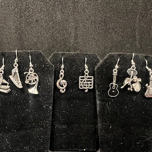 May include: A set of silver earrings featuring musical instrument and music note charms. The charms include a gramophone, harp, french horn, treble clef, music staff, guitar, drum set, and electric guitar.