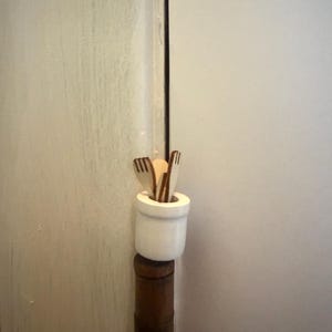 May include: A small, white ceramic container holding several wooden forks and a small, round object. The container sits atop a dark brown, cylindrical base, likely a decorative element. The background is a light-colored wall.
