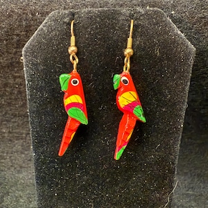 Puede incluir: Un par de pendientes con forma de loro. Los pendientes están pintados a mano con detalles rojos, amarillos y verdes. La cabeza del loro es verde y el cuerpo es rojo con detalles amarillos y verdes. Los pendientes cuelgan de ganchos dorados.