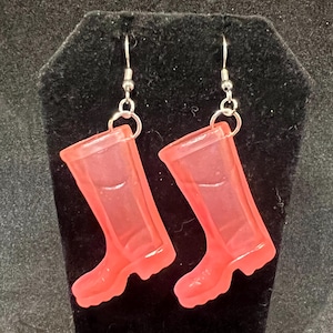 May include: A pair of pink rubber boot earrings. The earrings are made of translucent pink plastic and have a silver hook.
