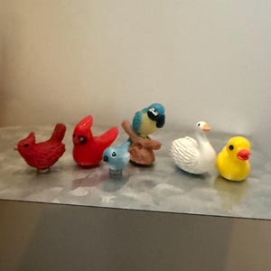 May include: A selection of small bird ornaments, featuring red cardinals, a blue bird, a parrot, a white swan, and a yellow duck. The figurines are made of a glossy material and are arranged in a line.