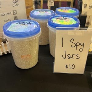 May include: Four clear plastic jars with blue lids filled with small, light-colored objects. Each jar has a colorful label that reads "I Spy Jar." A sign reads "I Spy Jars" and "$10."