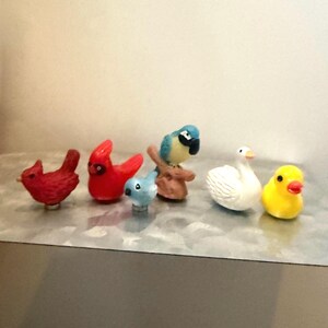 May include: A collection of six small bird figurines including two red cardinals, a tiny light blue bird, a blue and yellow parrot perched on a branch, a white swan, and a small yellow duckling, all arranged in a horizontal row on a flat surface.