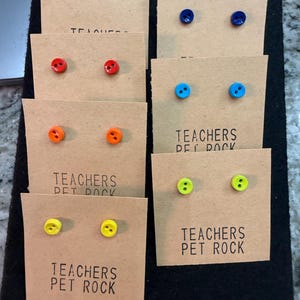 May include: Button stud earrings in various colors, including pink, red, orange, blue, and yellow. Each pair is mounted on a small, tan card with the text "TEACHERS PET ROCK" printed in black. The earrings are arranged on a black surface.