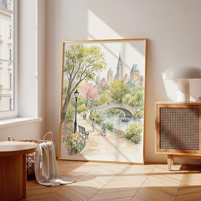 May include: A framed watercolor painting depicting a park scene with a stone bridge over a river, lush green trees, pink flowering bushes, a lamp post, a bench, and people walking and cycling along a path, with city buildings in the background.