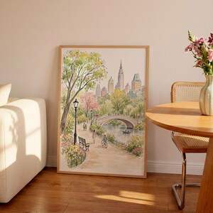 May include: A framed watercolor print depicting a park scene with a stone bridge over a body of water, a path with a black lamp post, a bench, and people walking or cycling, set against a backdrop of city skyscrapers in shades of green and pink.