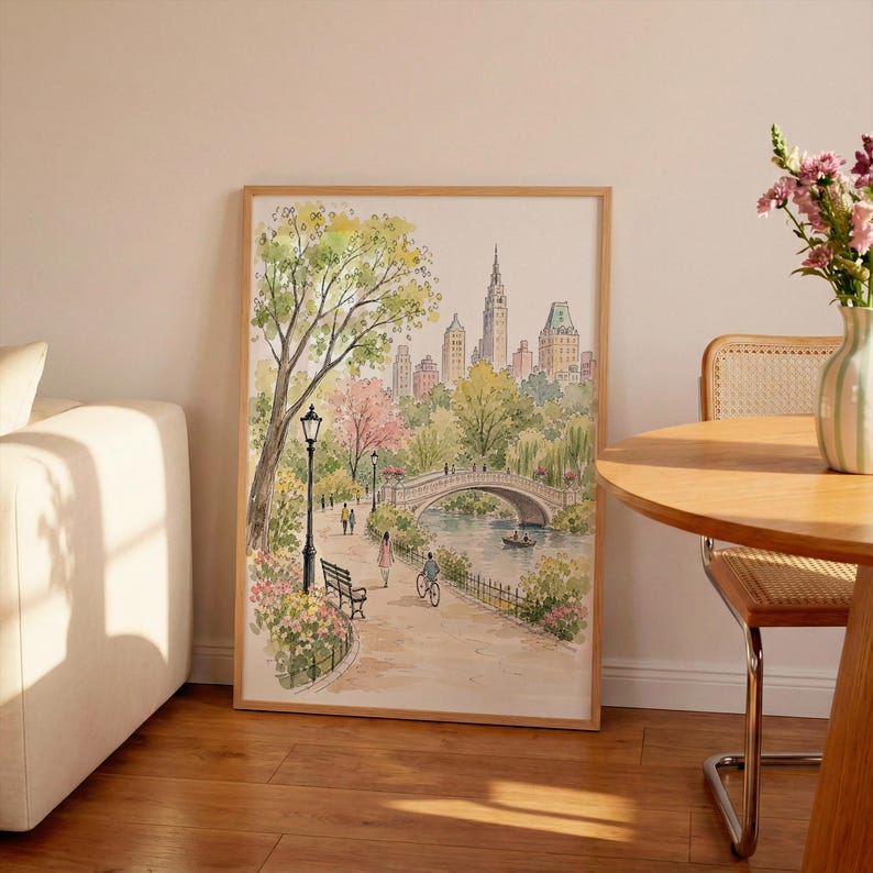 May include: A framed watercolor print depicting a park scene with a stone bridge over a body of water, a path with a black lamp post, a bench, and people walking or cycling, set against a backdrop of city skyscrapers in shades of green and pink.