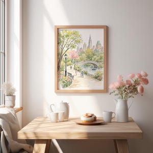 May include: A framed watercolor print depicting a park scene with a stone bridge over a pond, lush green trees, a lamppost, and city buildings in the background. The artwork is displayed on a wall above a wooden table with a white pitcher and pastries.