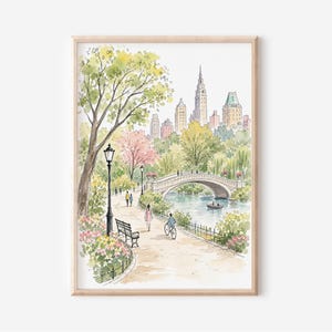 May include: A framed watercolor print depicting a park scene with a stone bridge over a pond, a winding path, a black lamppost, a bench, and people walking or cycling. In the background, a city skyline rises above lush green and pink trees.