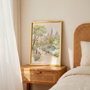 May include: A framed watercolor print depicting a park scene with a stone bridge over a body of water, surrounded by lush green trees, pink flowering bushes, and city buildings in the background. A path features a black lamp post and figures.
