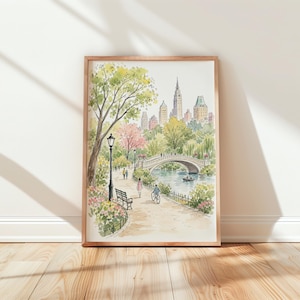 May include: A watercolor print in a wooden frame depicting a park scene with a stone bridge over a pond, a path with a bench, a lamppost, and people walking and cycling. In the background, a city skyline with tall buildings rises above trees.