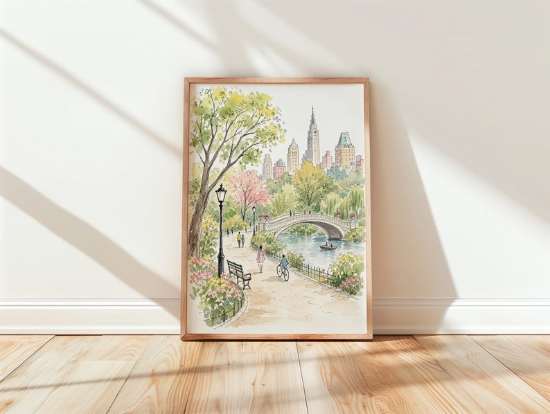 May include: A watercolor print in a wooden frame depicting a park scene with a stone bridge over a pond, a path with a bench, a lamppost, and people walking and cycling. In the background, a city skyline with tall buildings rises above trees.
