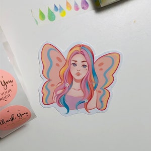 May include: A sticker featuring a colorful illustration of a girl with rainbow hair and butterfly wings. The girl is wearing a pink top and has white flowers in her hair. The sticker is on a white background.