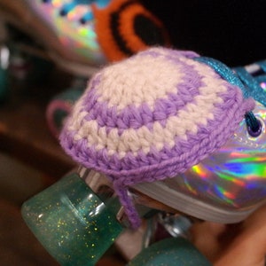 Könnte beinhalten: Nahaufnahme eines Rollschuhs mit einer gehäkelten Zehenkappe. Die Kappe ist weiß und lila. Der Schuh hat ein holografisches Finish, türkisfarbene Rollen und türkisfarbene Schnürsenkel.