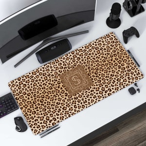 May include: A brown and tan leopard print desk mat with a monogrammed initial "S" in a gold frame. The mat is on a white desk with a computer monitor, a speaker, a microphone, a game controller, and a pair of headphones.