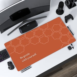 Peut inclure: Un tapis de bureau orange avec un motif de nuages blancs et le texte "Be gentle with yourself." Le tapis rectangulaire est conçu pour protéger une surface de bureau. Des accessoires informatiques entourent le tapis.