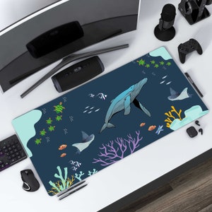 Puede incluir: Un ratón de ordenador azul y verde con temática oceánica con una ballena, tortugas marinas y coral. El ratón está sobre un escritorio blanco con un monitor de ordenador, un altavoz, un micrófono, un mando de juego y otros accesorios.