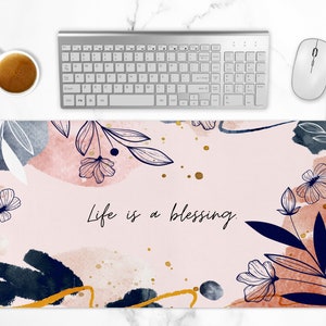 Puede incluir: Un tapete de escritorio de diseño floral rosa y blanco con el texto "Life is a blessing." en escritura negra. El tapete presenta formas abstractas de acuarela y detalles dorados.