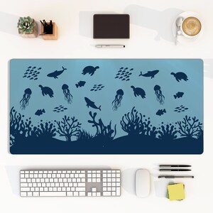 Puede incluir: Alfombrilla de escritorio azul con un diseño submarino que presenta siluetas de peces, ballenas, medusas y tortugas marinas. El diseño incluye corales y algas marinas en la parte inferior. La alfombrilla rectangular está sobre un escritorio blanco.