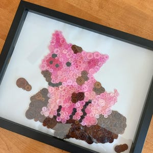 May include: Framed artwork depicting a pig, created from pink and brown buttons. The buttons are arranged on a white background within a black frame. A unique, textured piece of art, suitable for home decor.
