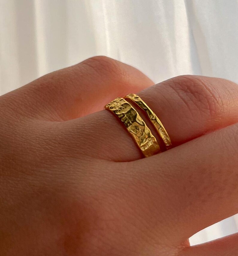 Pu&ograve; includere: Due anelli d'oro con una finitura testurizzata. Gli anelli sono impilati uno sopra l'altro su un dito.