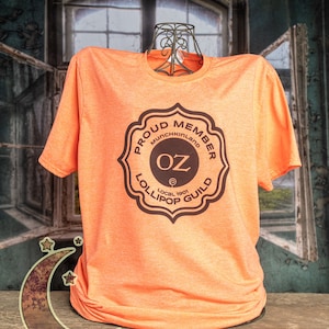 May include: Orange t-shirt with a brown graphic of a badge that says "Proud Member Munchkinland Oz Lollipop Guild Local 1901"