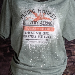 Peut inclure: T-shirt vert avec un motif de projection blanche. Le t-shirt présente un graphique d'un singe volant avec le texte "Flying Monkey Delivery Service" et "Just one cackle from the Wicked Witch. And we will come and carry you away. Your little dog too."