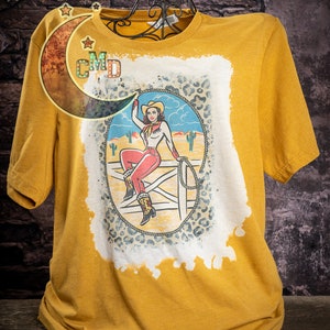May include: A mustard yellow t-shirt with a bleached design featuring a woman in a red and white outfit riding a horse. The design includes a blue sky, cacti, and a leopard print background.