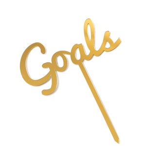 May include: Gold-colored metal cake topper with the word "Goals" written in a cursive font.