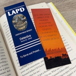 May include: Three bookmarks featuring a police badge, a cityscape, and a quote from the Harry Bosch series. The police badge bookmark is blue and gold with the text "Los Angeles Police Department LAPD Detective Hieronymus Bosch Serial No. 19558" and the text "To Serve and Protect..." The cityscape bookmark is orange and features a cityscape silhouette with the text "You are under arrest for murder, attempted murder and general conspiracy to be an asshole. - Harry Bosch" and the text "Michael Connelly". The quote bookmark is orange and features the text "You are under arrest for murder, attempted murder and general conspiracy to be an asshole. - Harry Bosch" and the text "Michael Connelly".