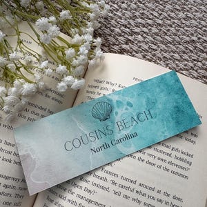 Könnte beinhalten: Ein Lesezeichen mit dem Text "COUSINS BEACH North Carolina" und einer Muschel-Illustration. Das Lesezeichen liegt in einem offenen Buch, mit weißen Blumen im Hintergrund. Das Lesezeichen hat ein blau-weißes Aquarelldesign.